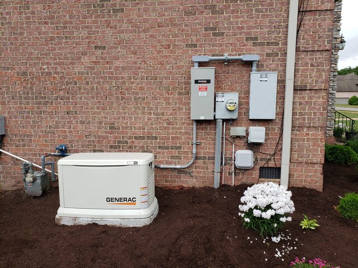 A Generac standby generator on its pad beside a brick home, with the transfer switch and meter on the wall behind it